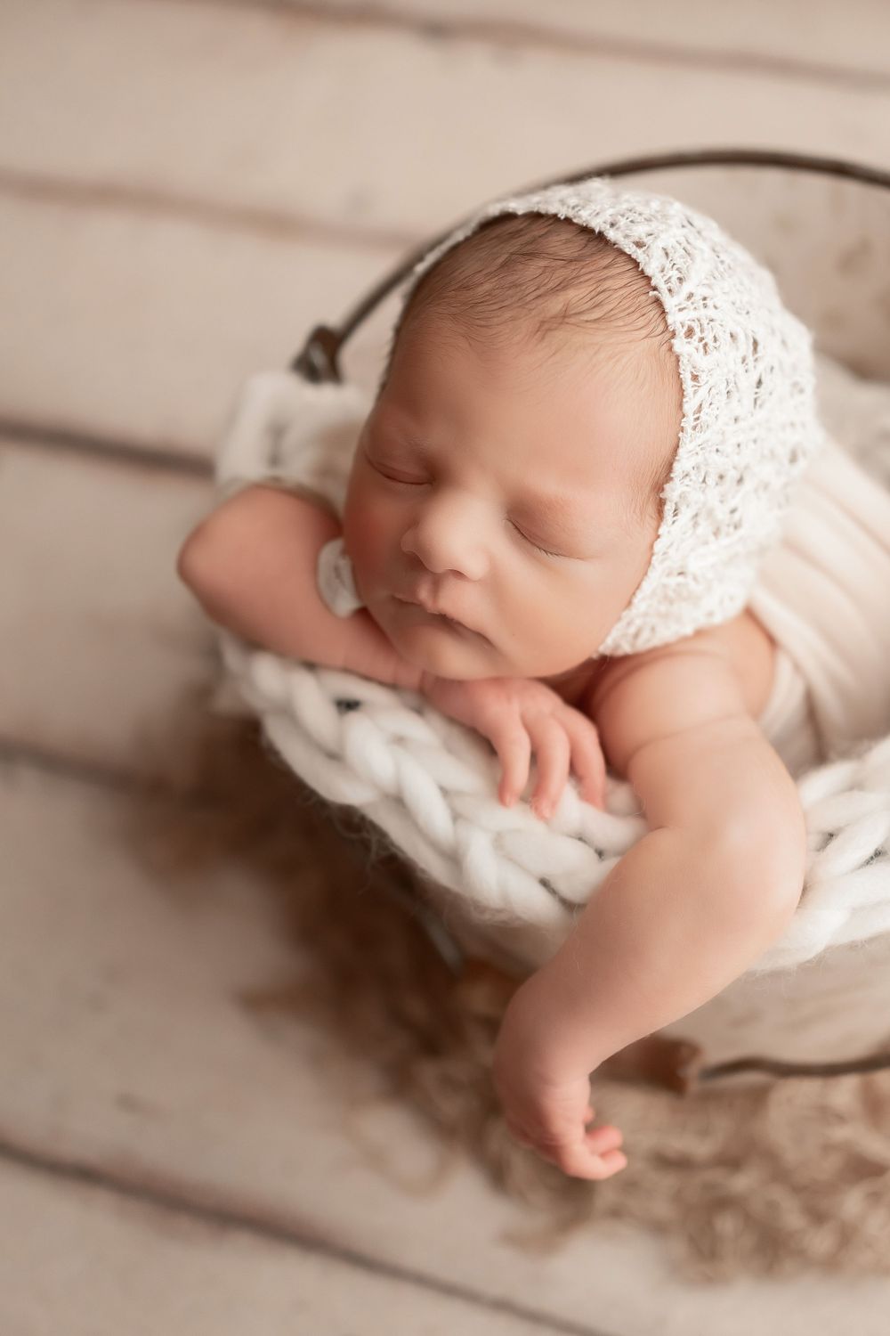 Sleeping baby wearing lace bonnet resting in knitted nest on wooden backdrop.