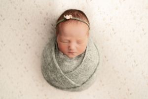 Sleeping newborn wrapped in sage green swaddle with dainty headband against white background.