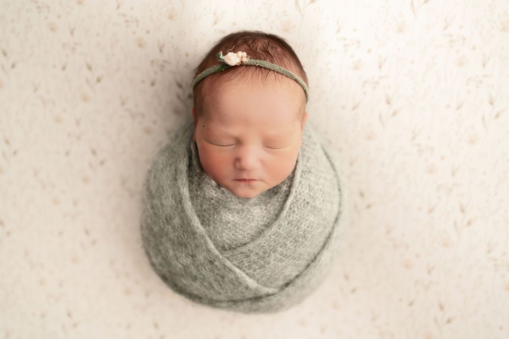 Sleeping newborn wrapped in sage green swaddle with dainty headband against white background.