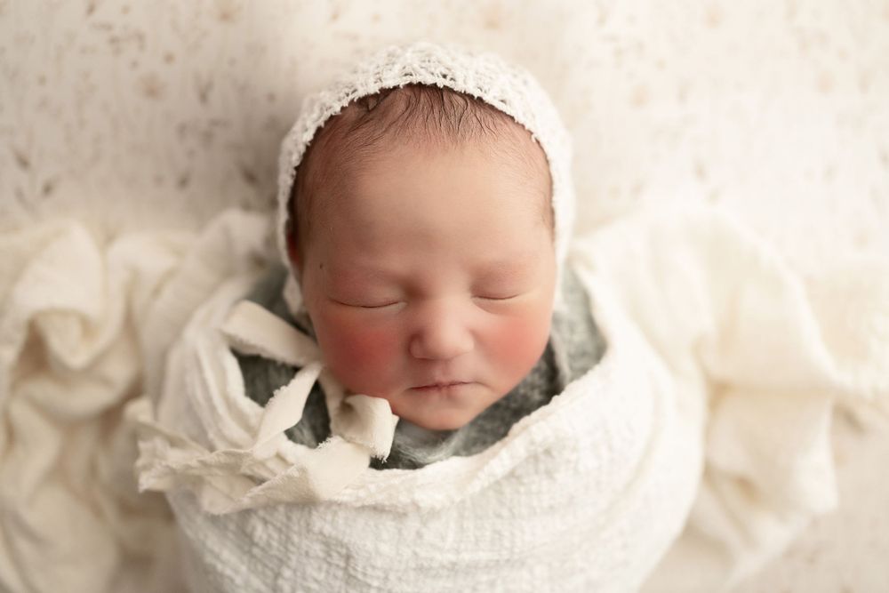 Sleeping newborn in white wrap and bonnet against cream textured background.