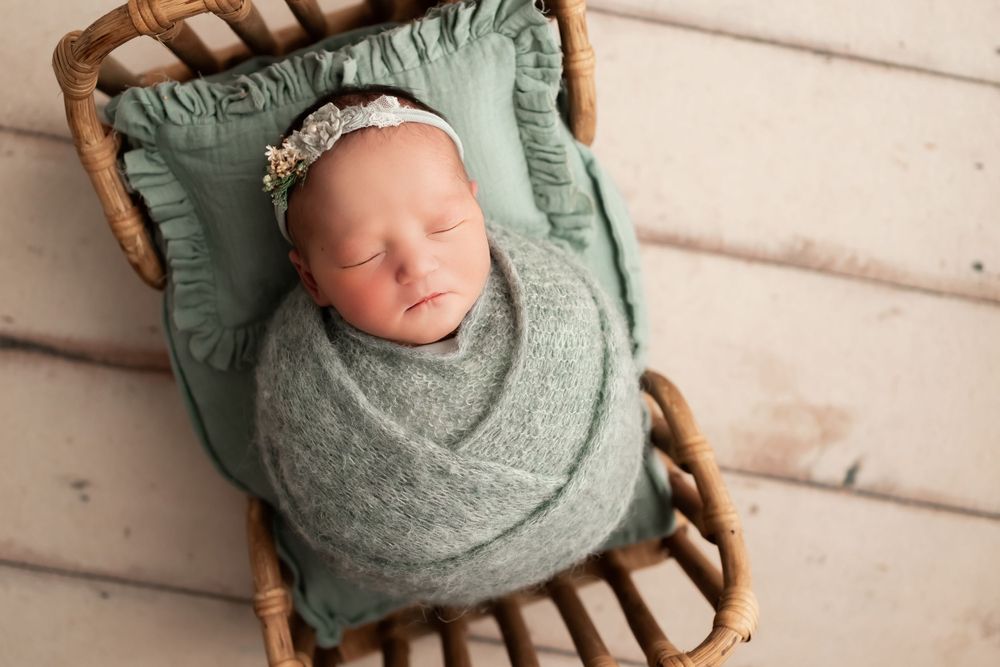 Newborn baby wrapped in sage swaddle sleeping on vintage wooden chair with mint cushion.