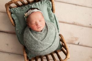 Newborn baby wrapped in sage swaddle sleeping on vintage wooden chair with mint cushion.