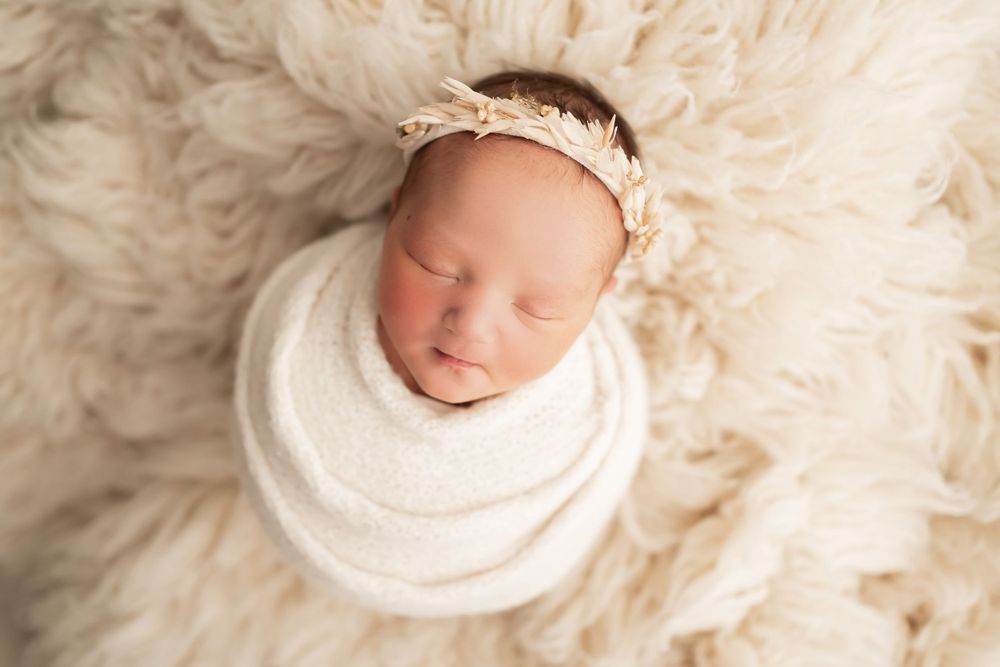 Peaceful newborn baby swaddled in white fabric on fluffy cream fur backdrop.
