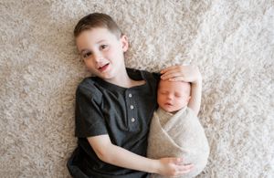 Siblings portrait with older child embracing newborn baby on white carpet.