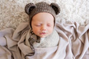 Sleeping baby wearing knitted bear hat swaddled in gray on white blanket.