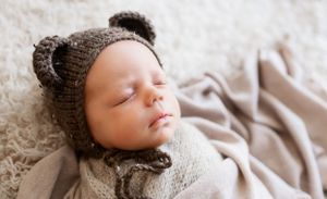 Sleeping newborn wearing a knitted brown bear hat on white textured blanket.