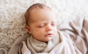 Peaceful sleeping newborn wrapped in soft gray swaddle on white textured surface.