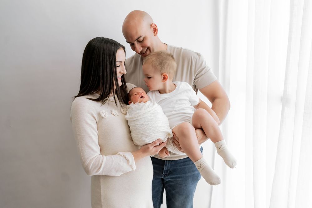 Parents in white and grey casual attire take turns holding and cuddling their newborn baby in front of a window.