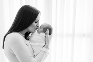 Loving parent gently kisses newborn baby wrapped in white swaddle against bright background.