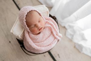 Peaceful newborn swaddled in soft pink wrap sleeping peacefully on white textured backdrop.