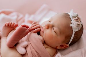 Sweet newborn baby in pink outfit rests on pink floral fabric with a delicate bow accessory.