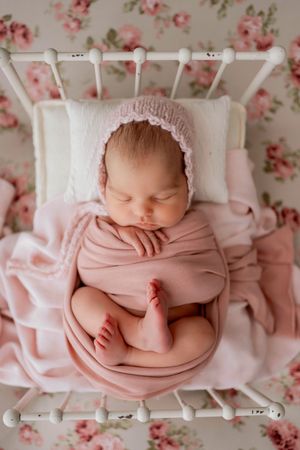Sleeping newborn wrapped in soft pink swaddle on vintage floral bedding with white headboard.