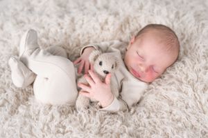 Newborn baby resting on soft white rug with small stuffed toy during professional photoshoot.