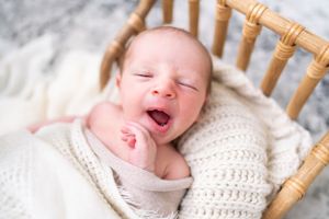 Newborn baby resting in wooden chair with knit wrap during professional portrait session.