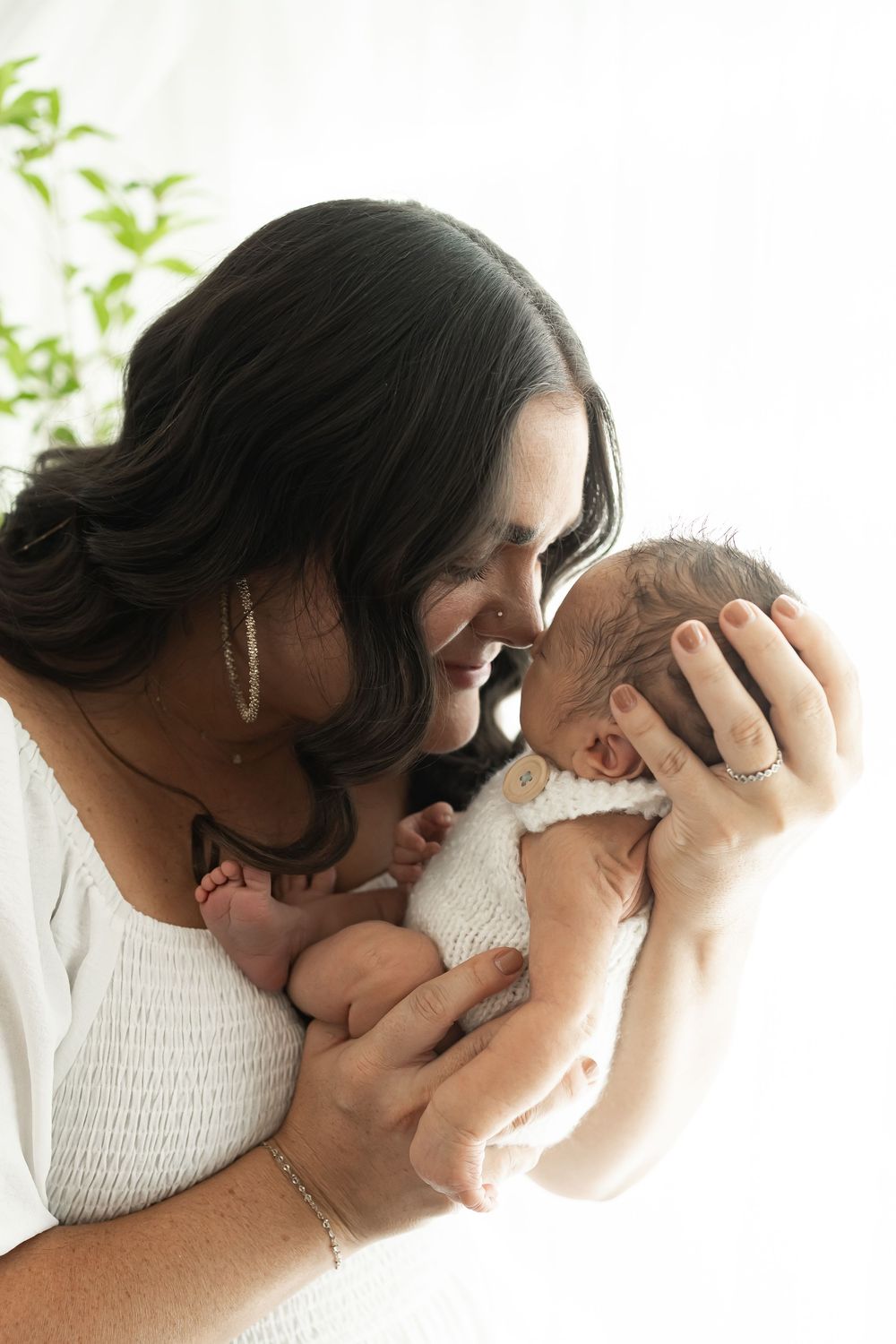 A smiling mother tenderly holds her newborn baby close to her face, dressed in white against a bright background.