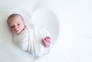 Baby wrapped in white swaddle lying on gentle white backdrop during newborn photography session.