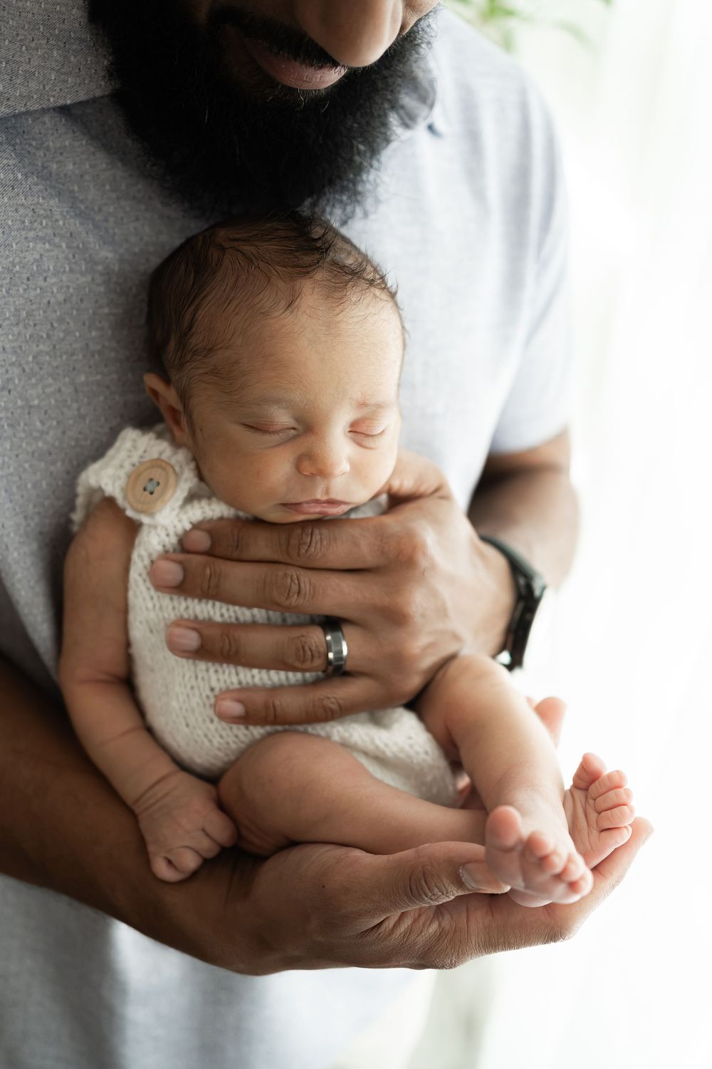 Bearded father tenderly cradling sleeping newborn baby dressed in white knit outfit, wearing a silver ring.
