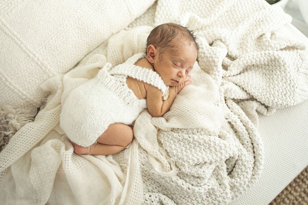 Newborn baby sleeping peacefully on white knit blanket wearing cream romper in soft natural light.