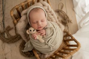 A sleeping newborn swaddled in soft beige fabric lies in a woven basket with a small teddy bear.