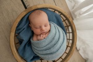 Small sleeping baby with blue swaddle blanket safely nestled in a round wooden bowl.