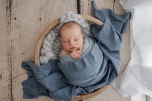 A sleeping newborn swaddled in blue knit fabric rests in a round basket on rustic wooden flooring.