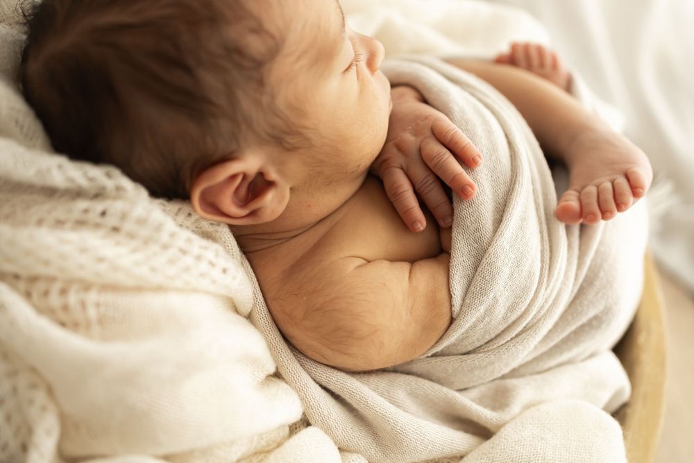 Peaceful newborn baby sleeping swaddled in soft beige wrap, curled up in cozy bowl with white knit blanket.