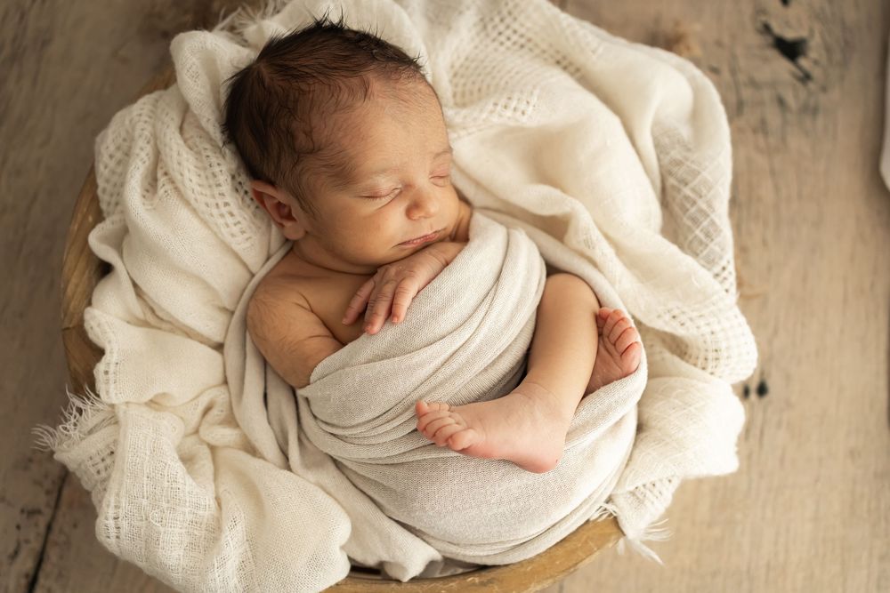 Newborn baby wrapped in cream white swaddle sleeping peacefully in a wooden bowl with soft blankets.