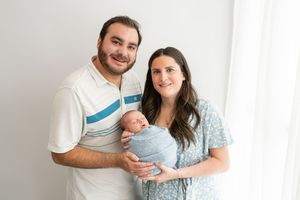 A couple holds a newborn baby wrapped in blue while standing against a white wall in casual attire.