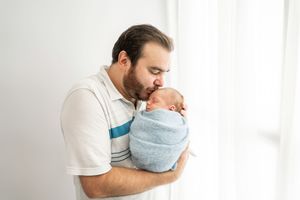 A tender moment between parent and swaddled newborn baby standing by white curtains in soft natural light.