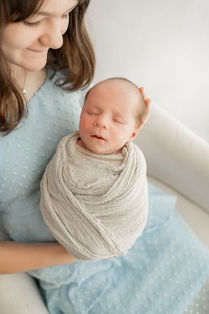 A sleeping newborn baby wrapped in a soft beige wrap is being held against a light blue patterned fabric.