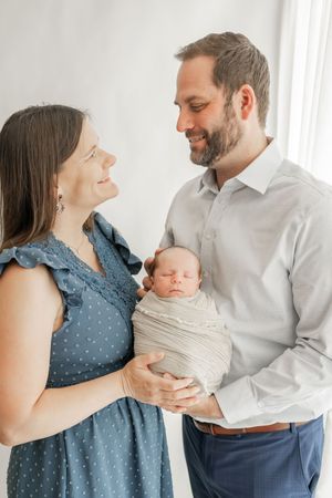 A couple in blue dress and white shirt share loving moments with their newborn baby in a bright indoor setting.