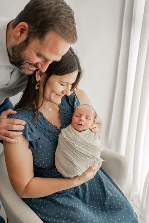 Parents share intimate moments with their newborn baby, dressed in white and blue against a bright window backdrop.