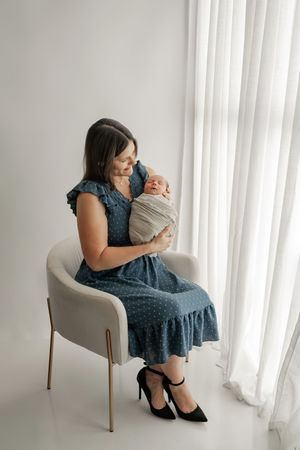 Collage of someone in blue dress sitting in beige accent chair holding baby against white curtained window.
