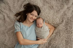 A tender moment captured showing a parent in light blue embracing a swaddled newborn against a textured wall background.