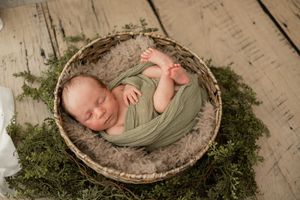 Newborn baby peacefully sleeping in a rustic basket surrounded by green moss and wrapped in sage green fabric.