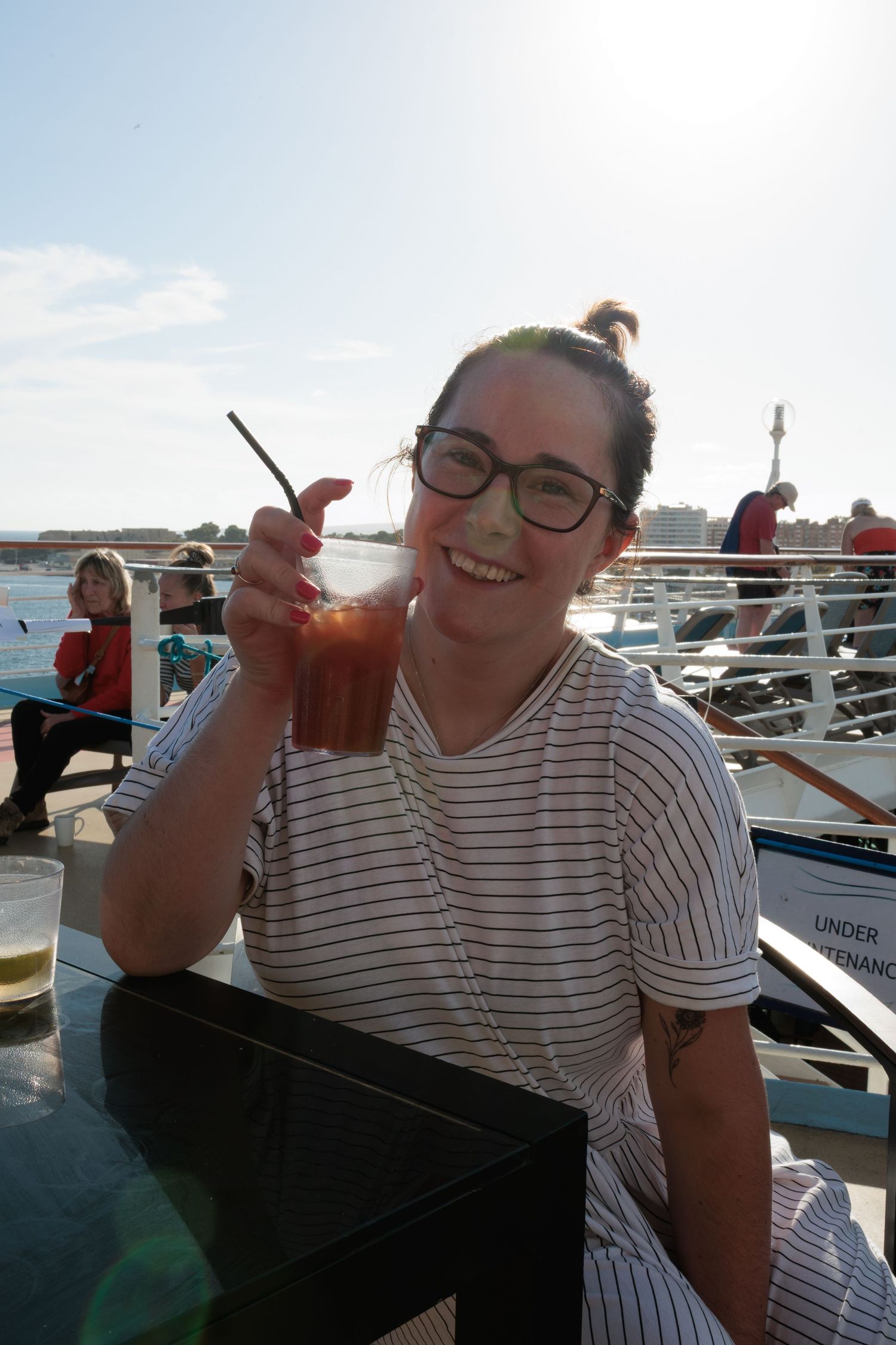 Smiling young woman with glasses enjoying a cocktail with a straw on a sunny boat deck.