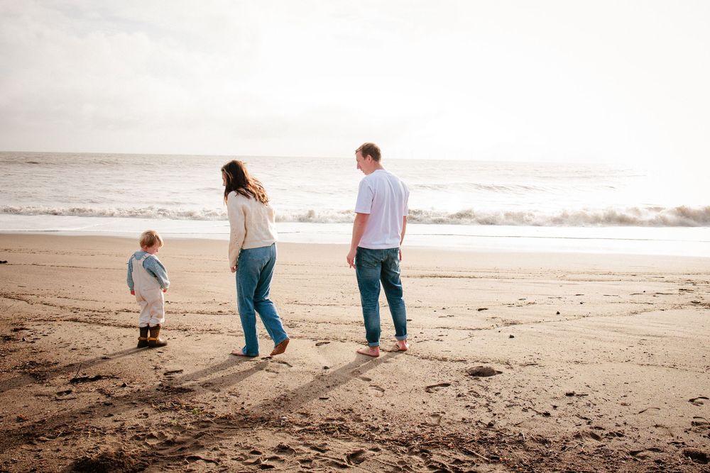 Young family with toddler walking along a sandy beach at sunset, viewed from behind.