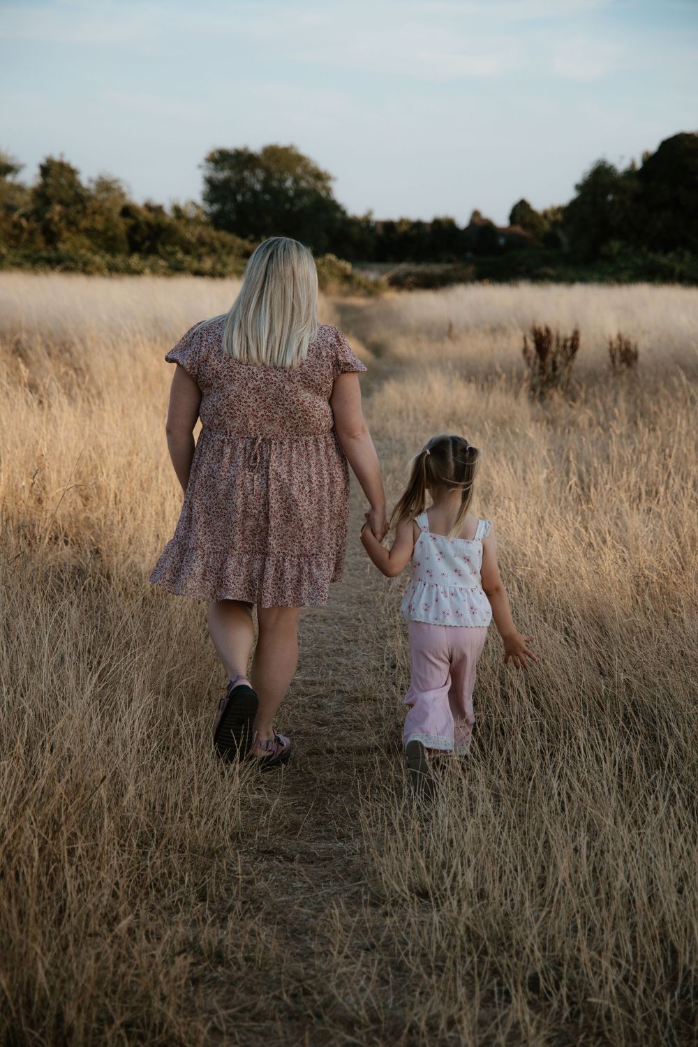 A woman and young girl walk hand in hand through a golden wheat field on a summer evening.