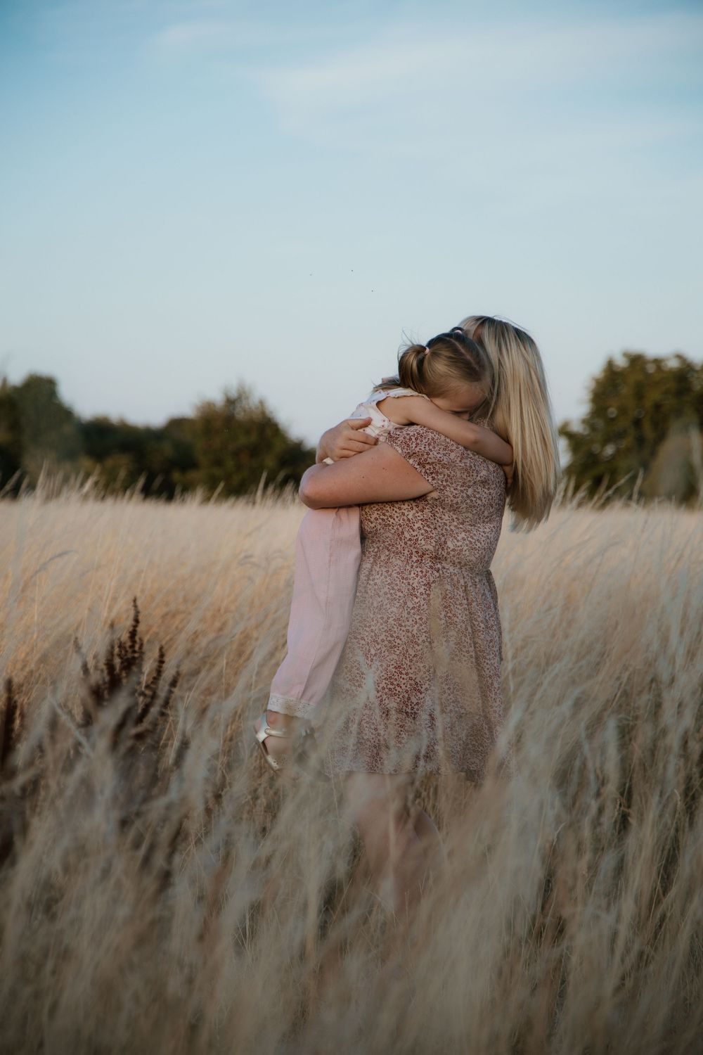 A mother and child share a tender embrace in a golden wheat field under a soft blue sky.