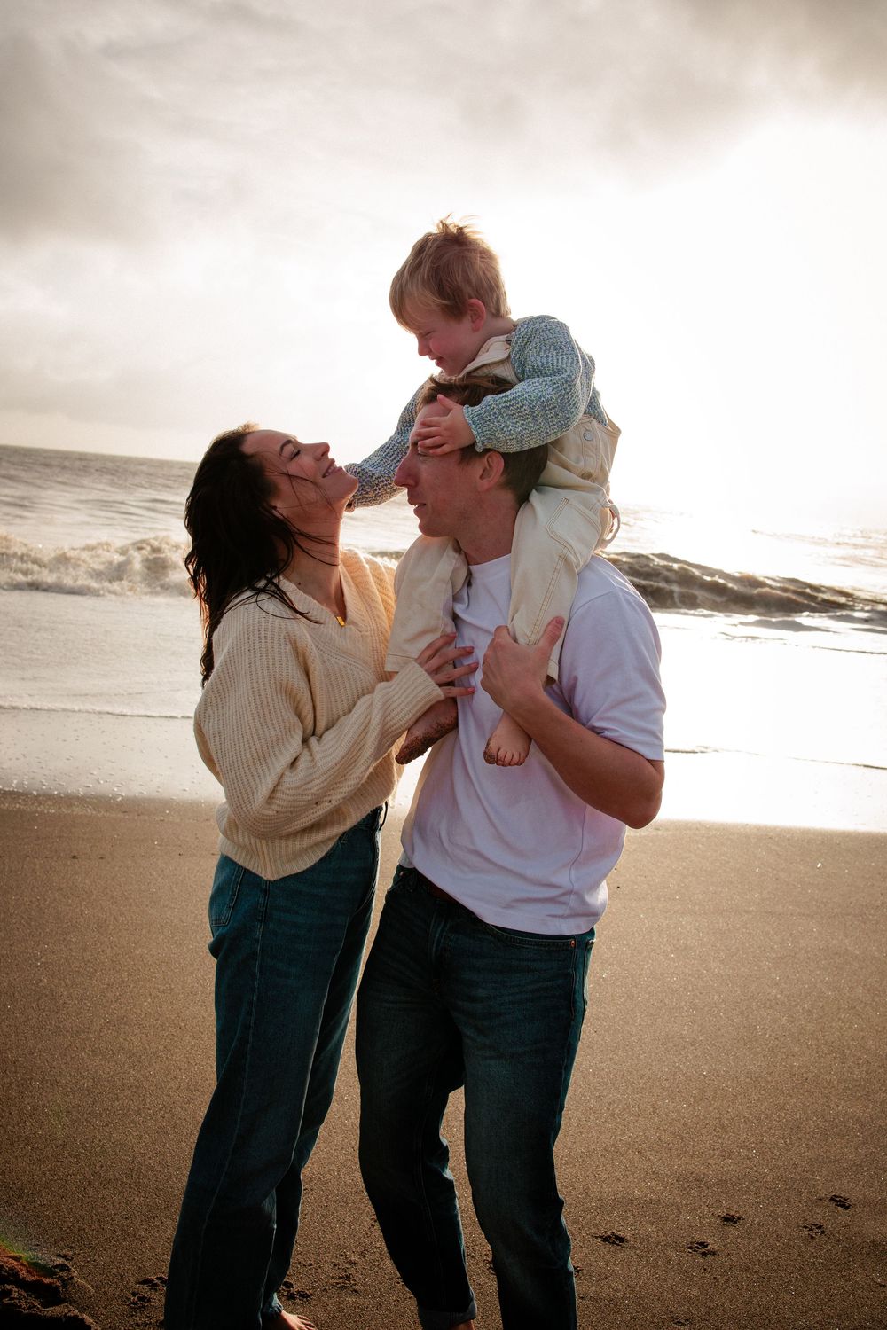 Joyful family at the beach with father carrying child on shoulders while mother laughs beside them.