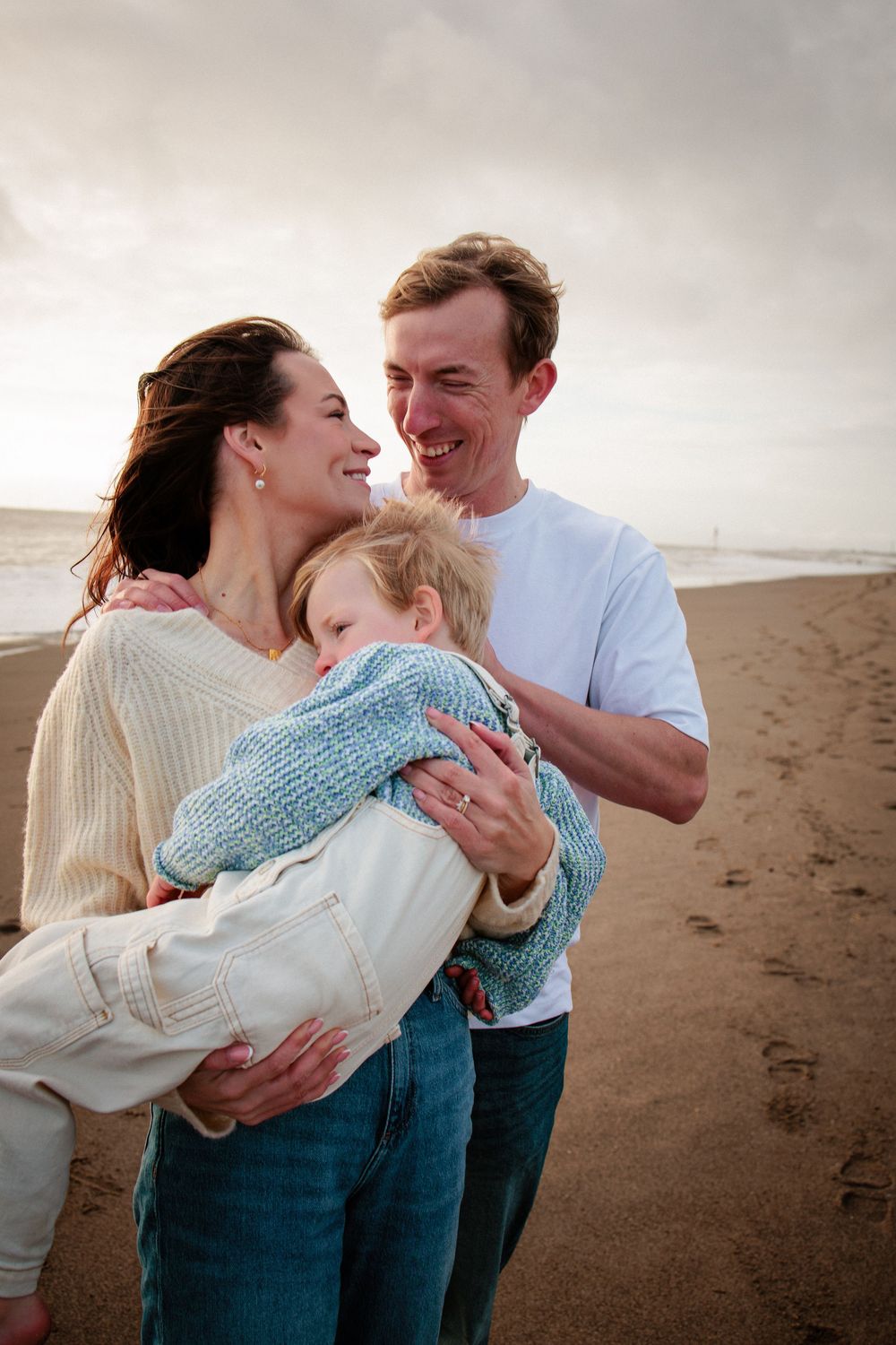 Smiling family of three embracing on a sandy beach with ocean waves in the background.