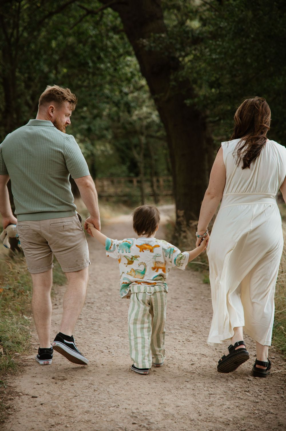 A family of three walks hand-in-hand down a forest path, with a toddler in a dinosaur outfit between two parents.