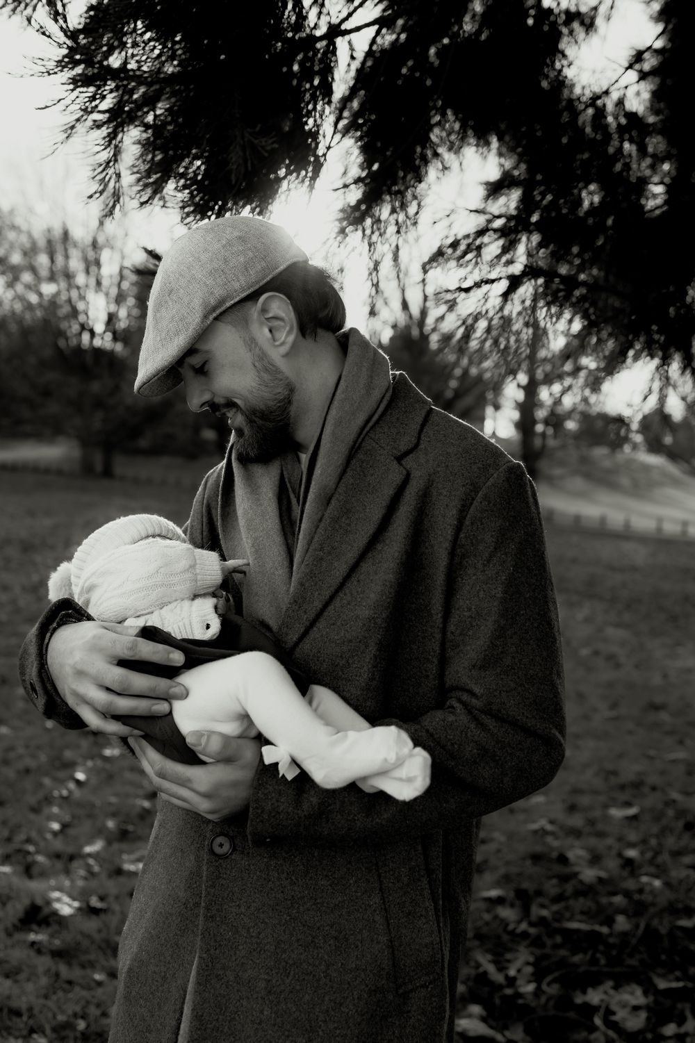 Black and white photo of a bearded father in a coat and hat tenderly holding his newborn baby outdoors.