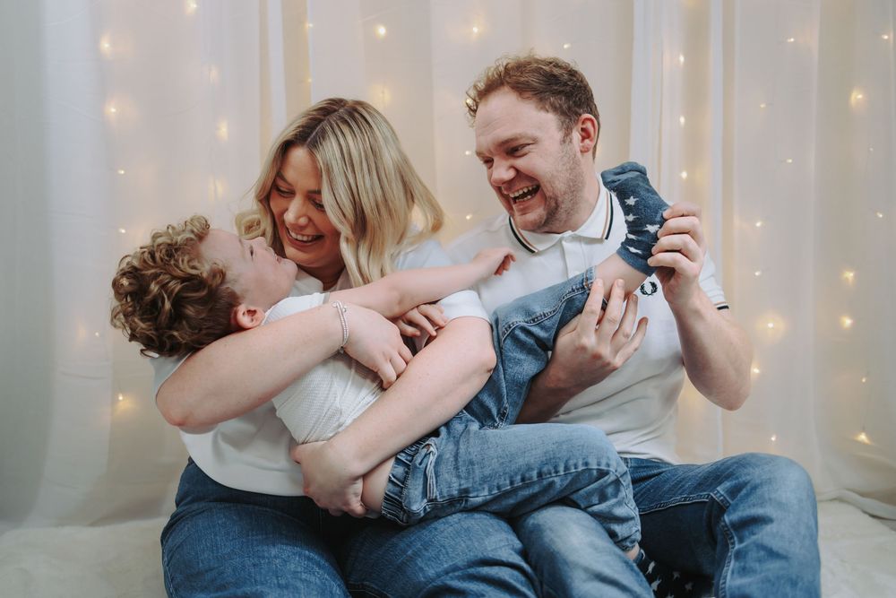 Happy family of three laughing and playing together on a bed with warm fairy lights in the background.