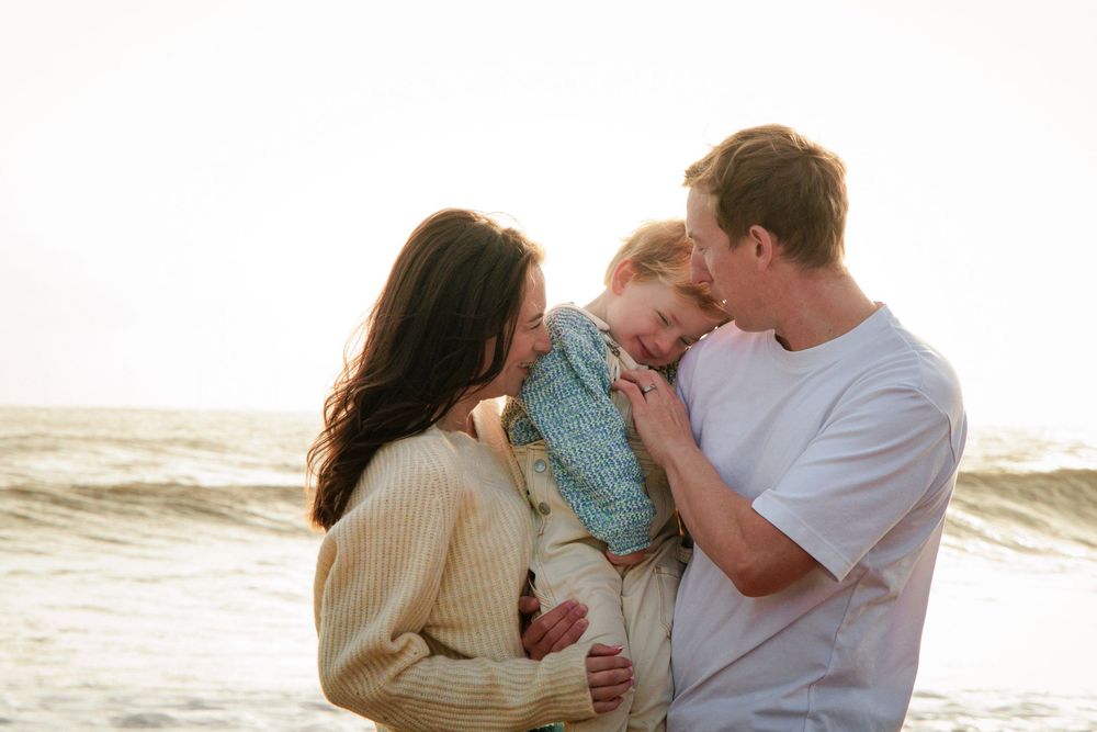 Loving parents holding their young child between them on a sunlit beach by the ocean.
