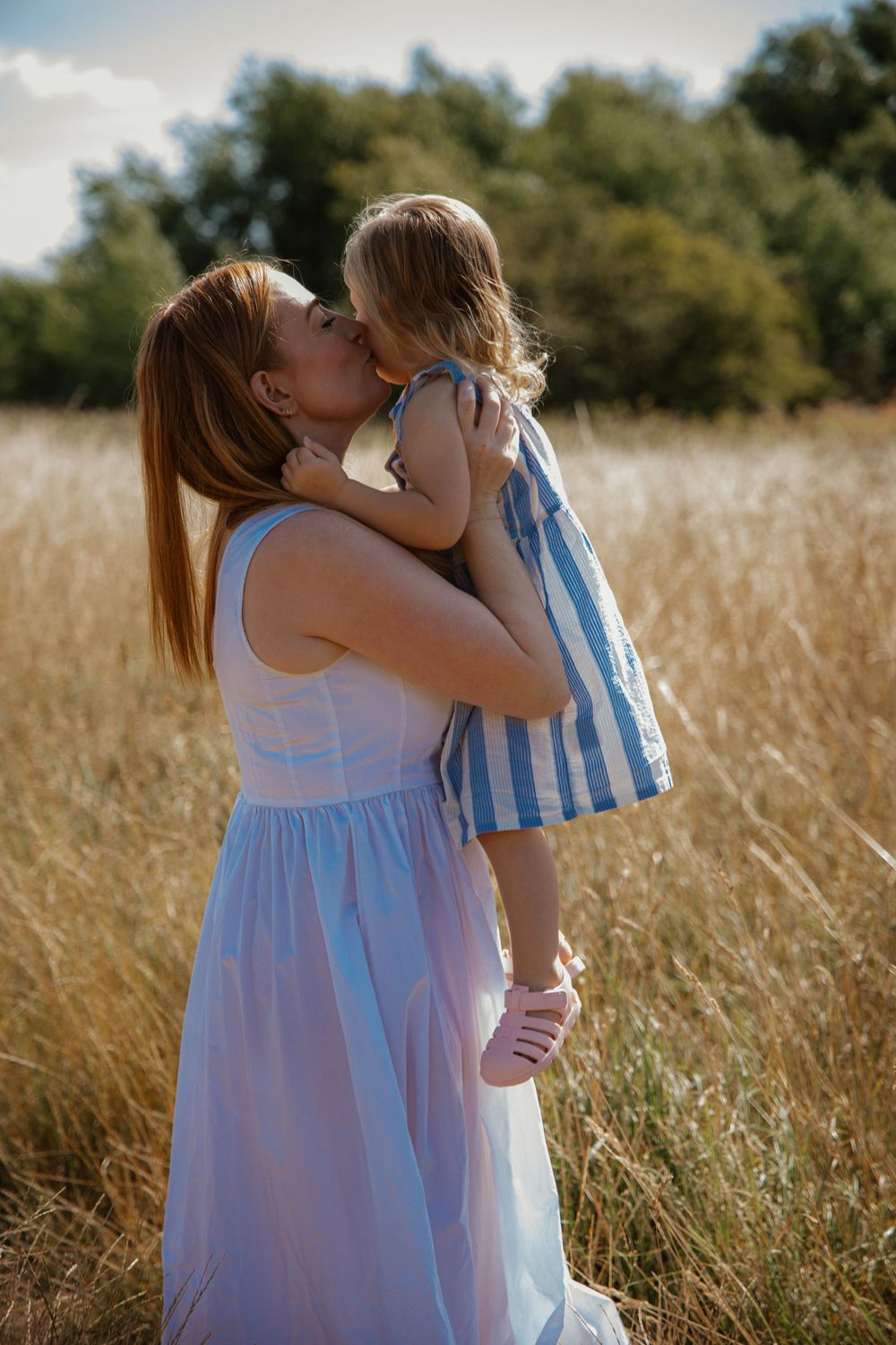 A mother in a blue dress kisses her young daughter in a striped dress while standing in a golden field.