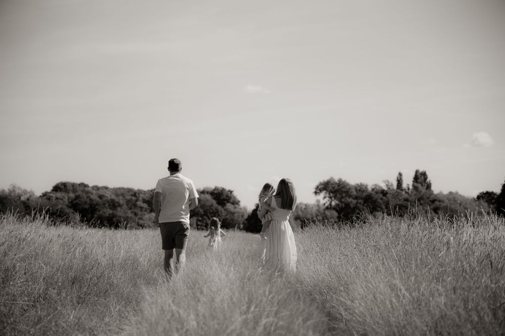 Black and white photo of a family walking through a field, parents holding child's hands amid tall grass.