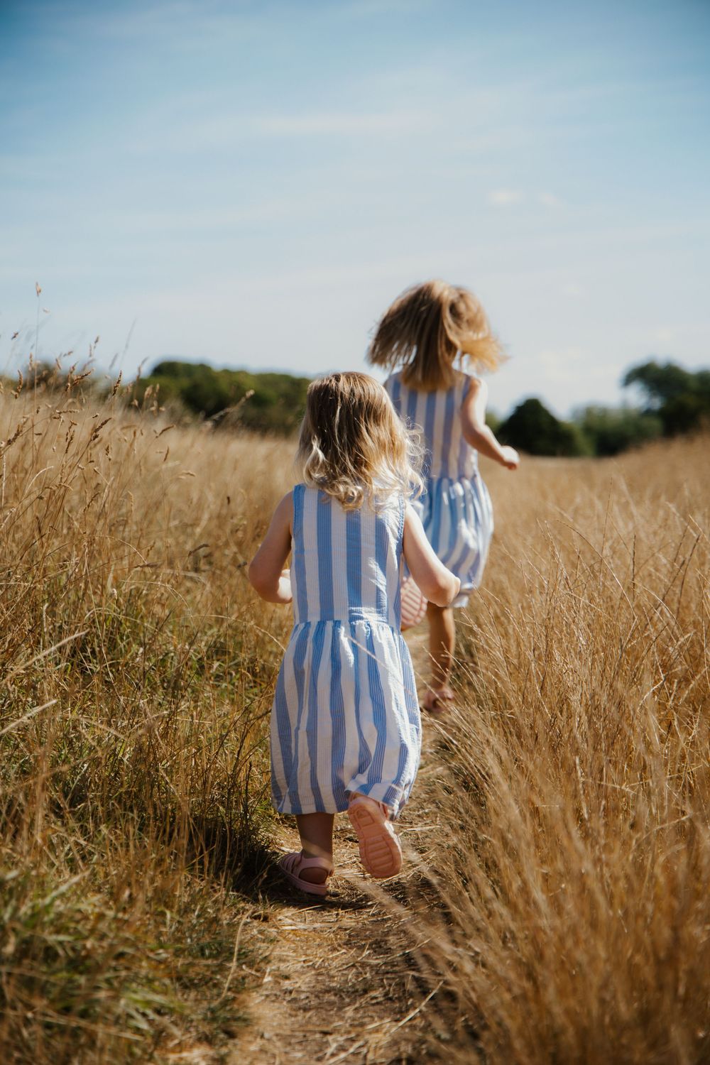 Two young girls in matching blue striped dresses running along a golden wheat field path on a sunny day.