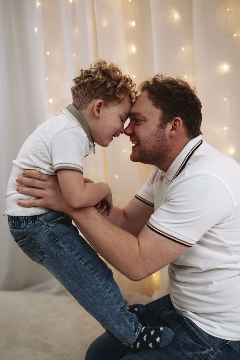 Loving father holding his curly-haired son close, touching foreheads and smiling in a warmly lit room.