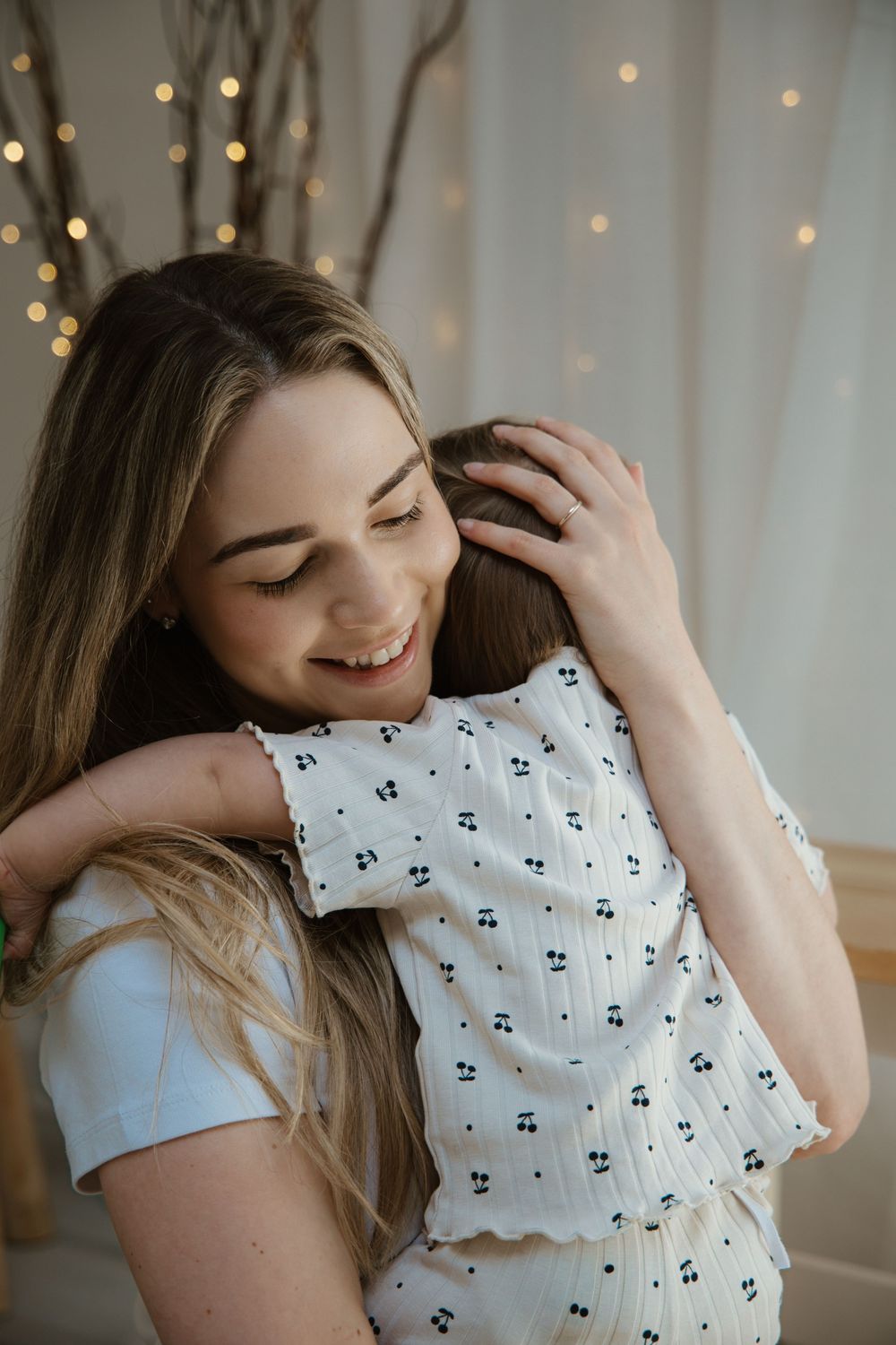 Smiling young woman with long blonde hair hugging a child in a white cherry-print outfit indoors.